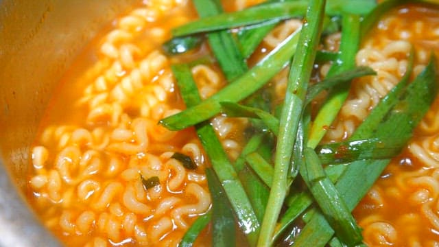 Bbowl of hot ramen topped with green onions steaming in a metal pot.