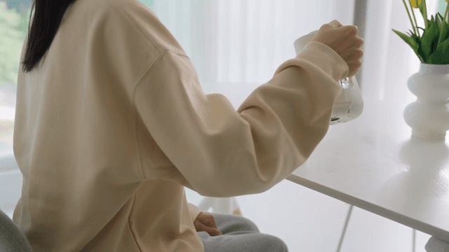 Woman pouring water into cup with coffee pot on table in bright living room