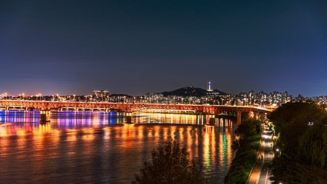 Colorfully lit bridge over a river at night with a cityscape in the background
