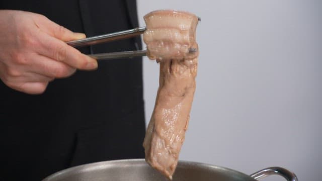 Person preparing boiled pork belly with tongs in a kitchen