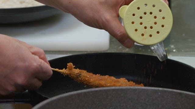 Adding seasoning to a fried food skewer in a pan