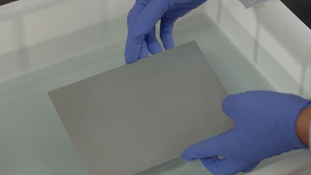 Gloved hands dipping a metal sheet into solution
