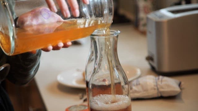 Pouring fermented kombucha into a glass jar on the kitchen counter