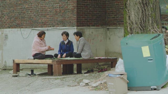 Three elderly women sitting and conversing on a wooden platform outdoors in a courtyard.