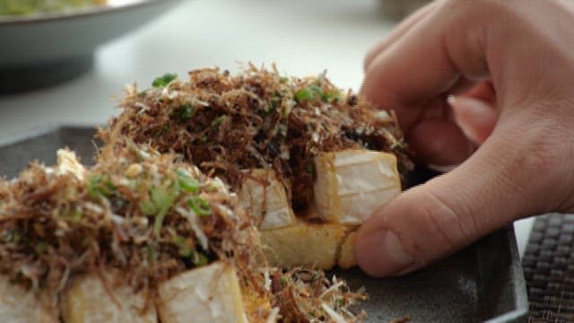 Hand Picking up an Appetizer Topped with Katsuobushi and Cheese