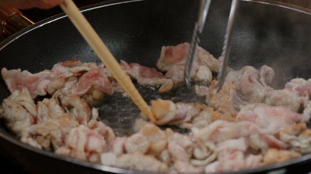 Pork being fried in an oiled frying pan with tongs and chopsticks