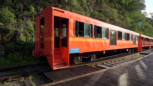 A bright red train waits on the tracks near a lush, green forest on a sunny day.