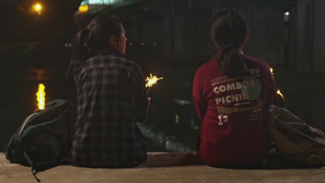 Two girls with sparklers sit by the water at night