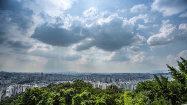 A panoramic view of a city skyline under a cloudy sky from a lush green hilltop.