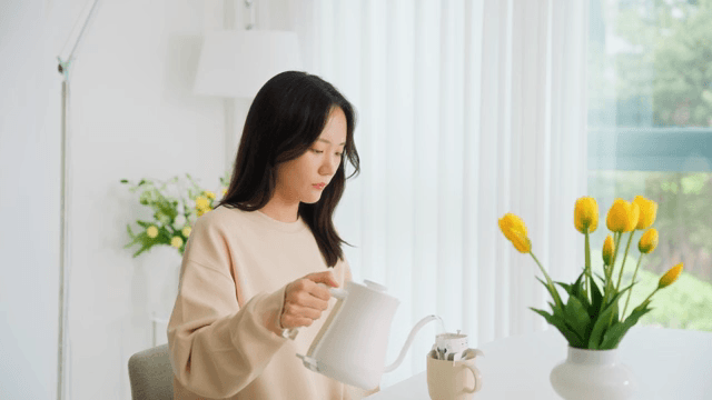 Woman pouring water into cup with coffee pot on table in bright living room