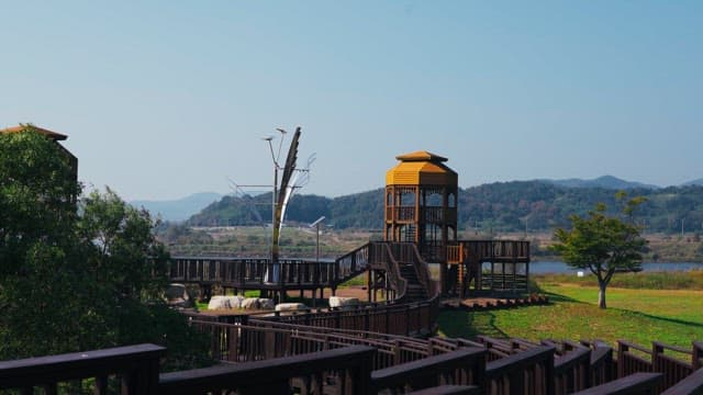 Scenic viewpoint with a yellow wooden tower and a modern sculpture, set by a river in a green park
