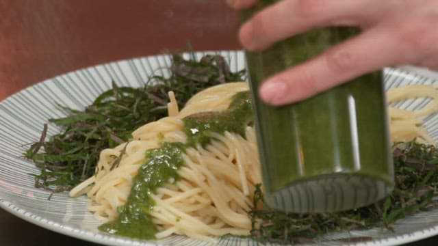 Person pouring green sauce on spaghetti with seaweed stems