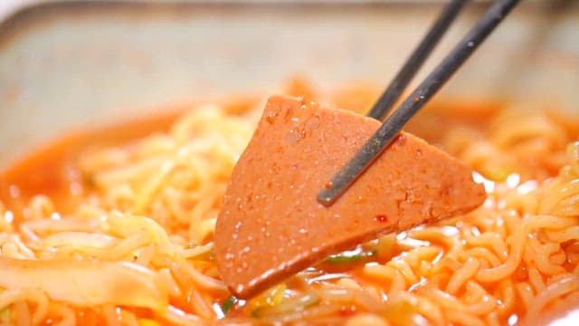 Chopsticks holding a slice of ham in ramen