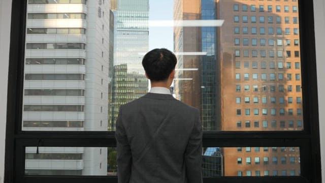 Man in a suit stretching while looking at the tall building outside his office window