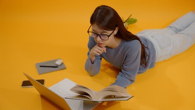 Woman with glasses studying with a laptop and a book on a yellow floor.