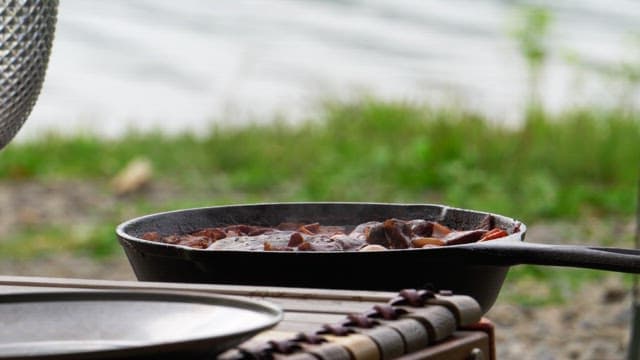 Person covering a frying pan with a lid while cooking food near a lake
