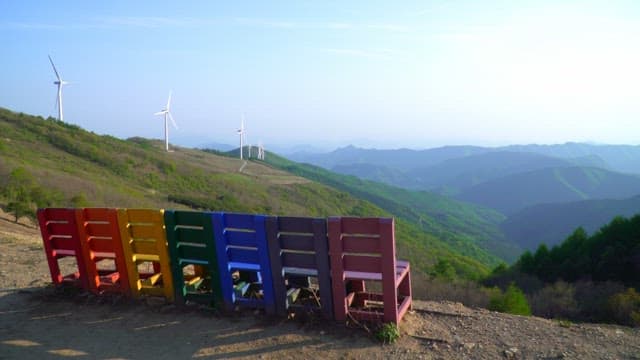 Rainbow-colored chairs displayed on top of a mountain with wind turbines in view