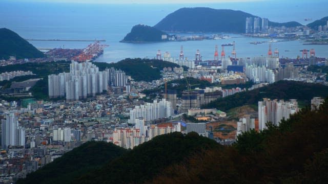Panoramic view of a coastal city surrounded by green mountains under a cloudy sky