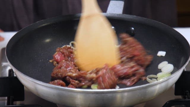 Stir-frying seasoned meat in a frying pan with a wooden spatula