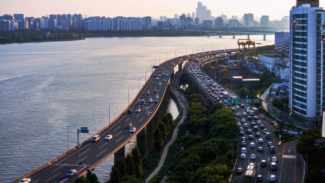 Vehicles on a highway by a river during sunset with a city skyline in the background