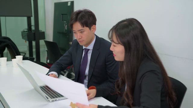 Two colleagues discussing documents and working on a laptop in an office setting