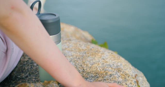 Person on a Rock Near the Water and Enjoying a Drink in Peace