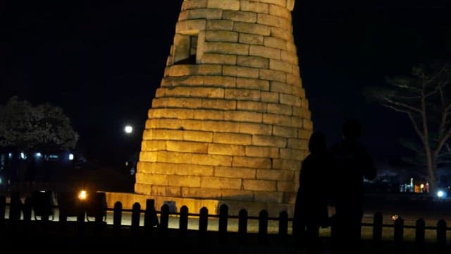 Two people walking with Cheomseongdae illuminated at night in the background