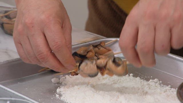 Hands preparing mussel skewers with flour coating in a kitchen