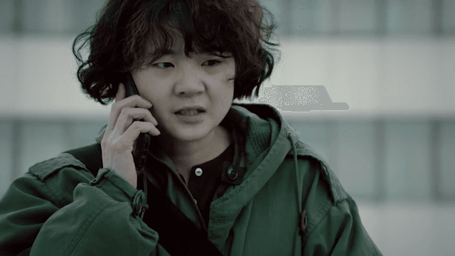 Person in a green jacket talking on a phone while standing outside in front of a building.