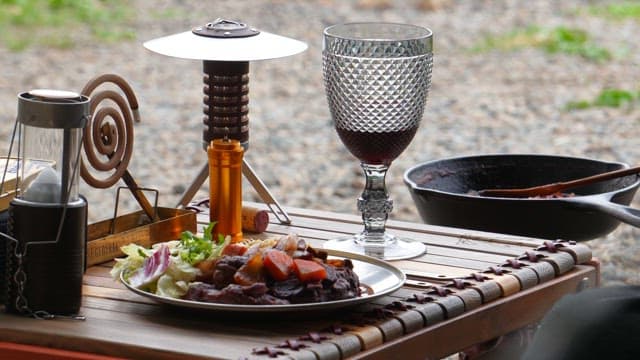 Outdoor meal setup with red wine, cooked meat and camping gear