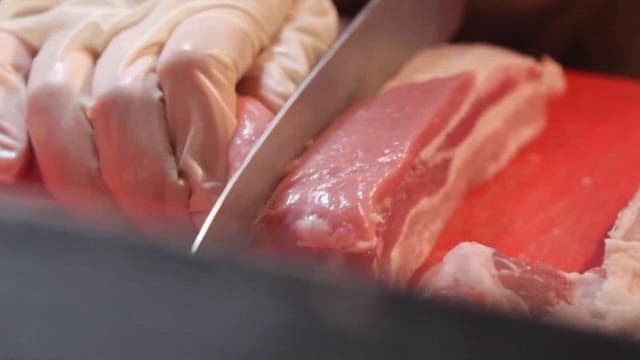 Person slicing fresh pork with a knife on a red cutting board