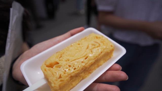 Hand Holding a Fresh, Golden Tamagoyaki in an Outdoor Market