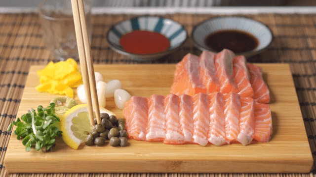 Scene of eating capers and pickled radish on a wooden cutting board with salmon sashimi on it