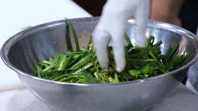 Gloved hand marinating chives in a stainless steel bowl