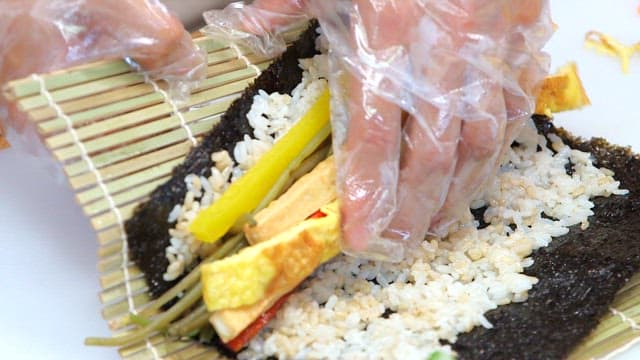 Gloved hands wrapping gimbap on a cutting board