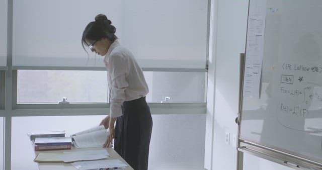 Woman organizing papers in a bright classroom next to a whiteboard.