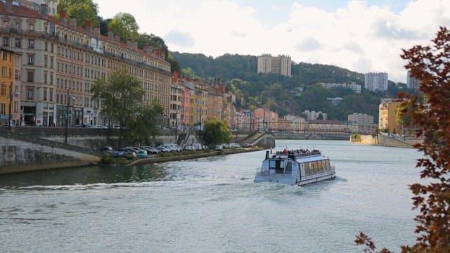 River cruise boat sailing through a European city with colorful buildings and a scenic hillside