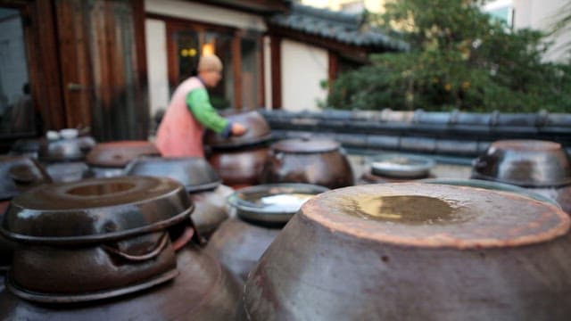 Woman in traditional Korean clothing checks large earthenware jars outside a wooden house.