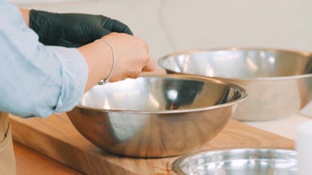 Person in gloves cracking an egg into a mixing bowl on a wooden counter